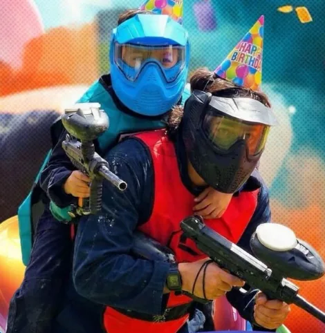 Dos jugadores con máscara y chaleco de paintball, uno con gorro de cumpleaños, frente a un fondo de globos