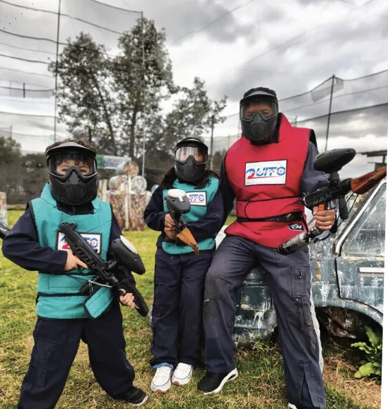 Dos niños y un adulto con máscara, chaleco y marcadora, listos para jugar paintball junto a una camioneta de la cancha