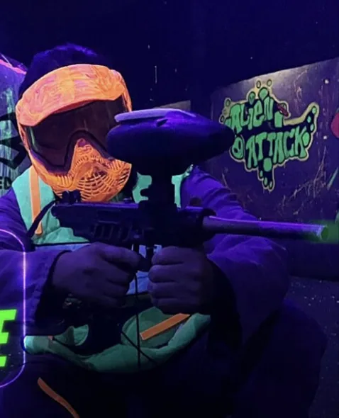 Jugador con máscara naranja fluorescente apunta su marcadora bajo luz ultravioleta, junto al grafiti Alien Attack